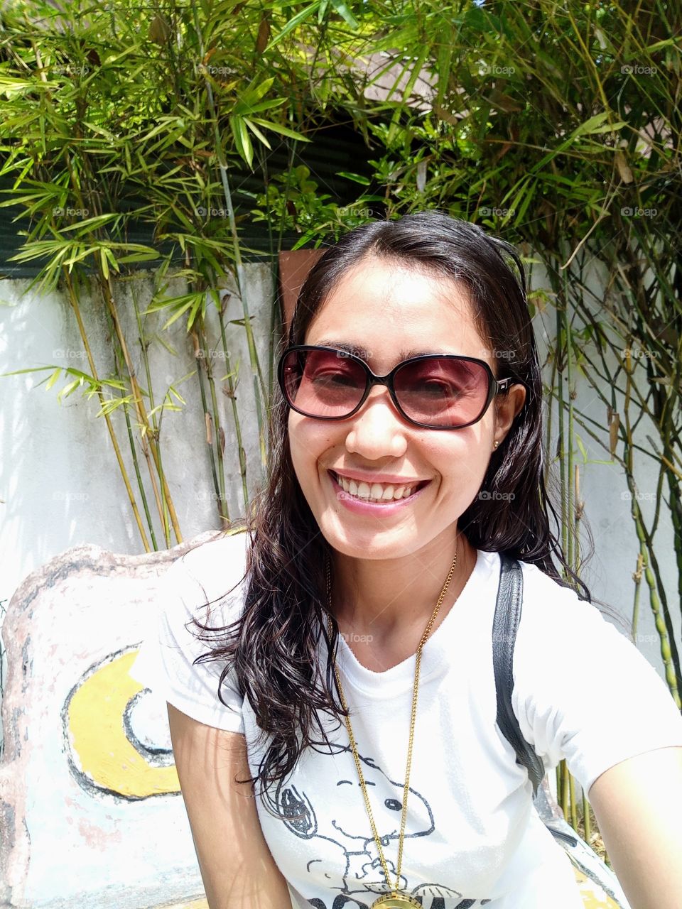 A woman smiling wearing sunglasses, white shirt, with bamboo and wall background. 