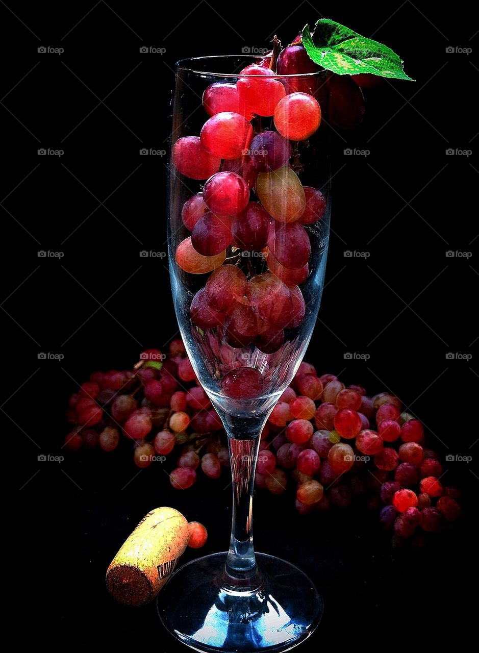 Fruit.  In the glass is a bunch of pink grapes with a green leaf.  Next to the glass is a bunch of pink grapes and a wooden cork from a bottle of wine.  Black background