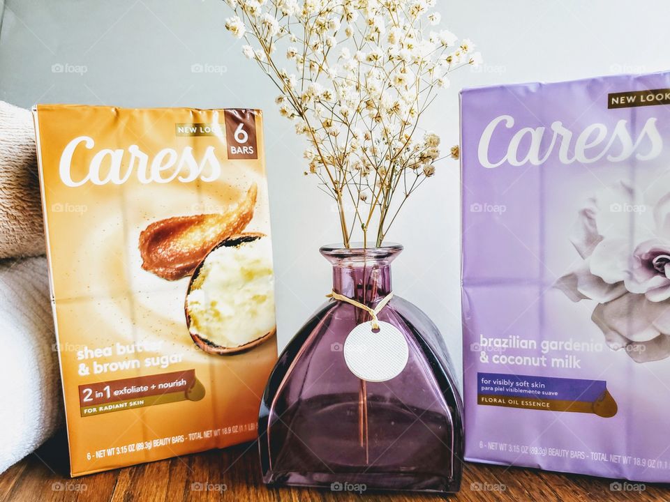 The new look Caress bar six pack of shea butter & brown sugar and brazillian gardenia & coconut milk bar soap. A purple vase with white flowers in the center. Towels on the left. On a wood surface.