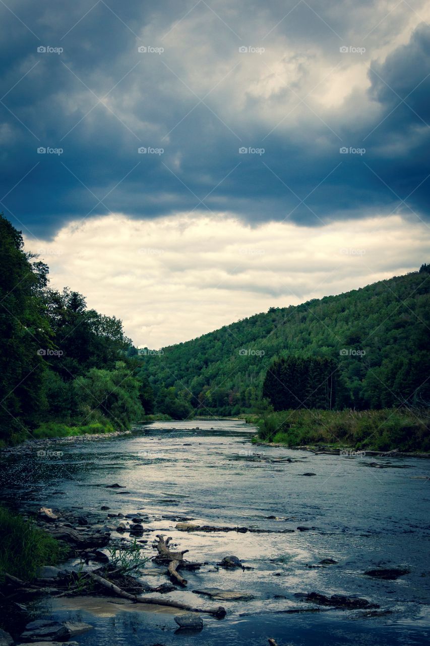 A moody and scenic portrait of a river in a forest between some mountains. with a moody and cloudy sky above it and the surrounding woods.