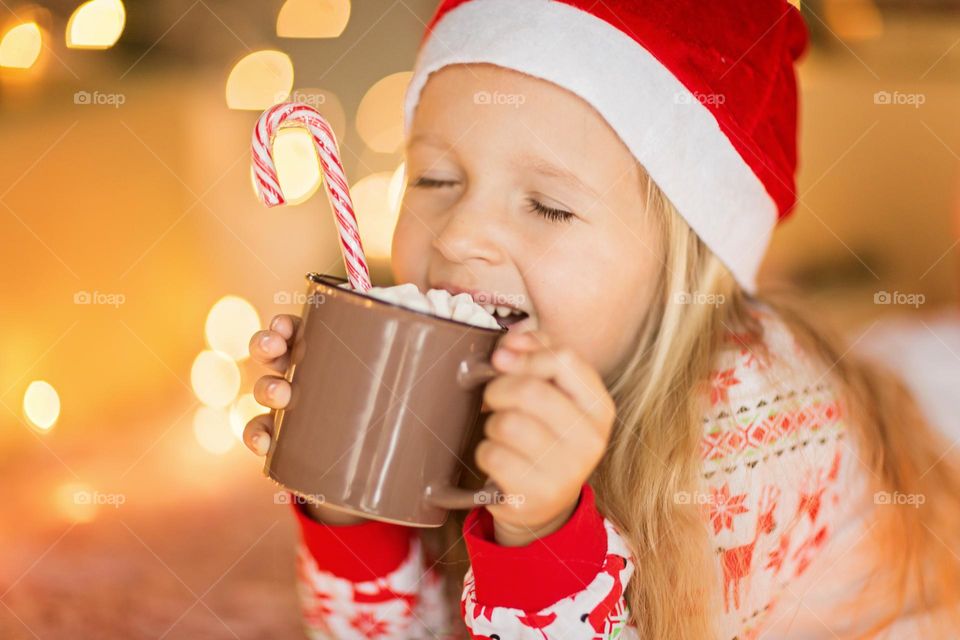 Little girl wearing Christmas pajamas and drinking hot cocoa with marshmallow at home 