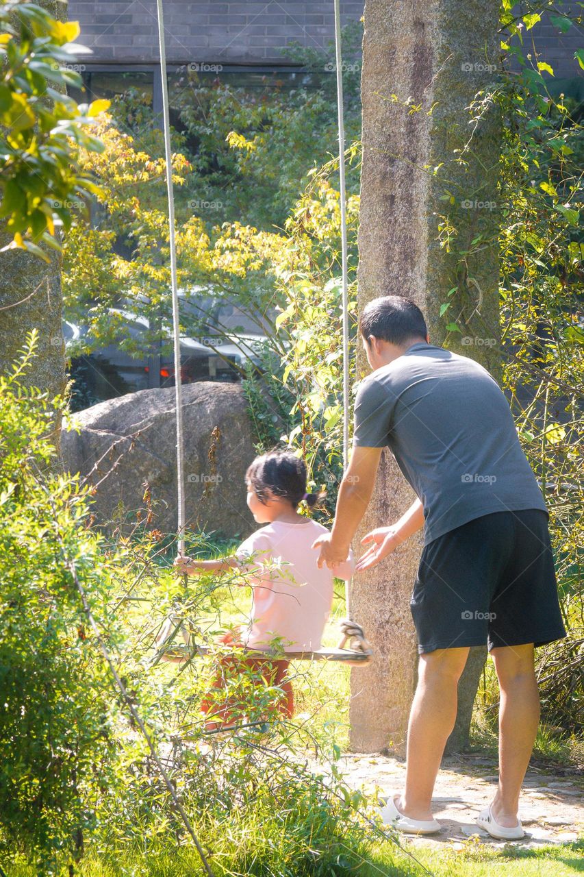 A daughter is swinging on a seesaw with her father in the park, creating a scene filled with love under the sunshine.