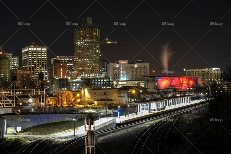 Foap, Day and Night: Nighttime skyline of Raleigh North Carolina as seen from Boylan Bridge. Scene includes the train station, the color-changing shimmer wall (currently red), and the orange crescent moon tucked away in a corner among the buildings.