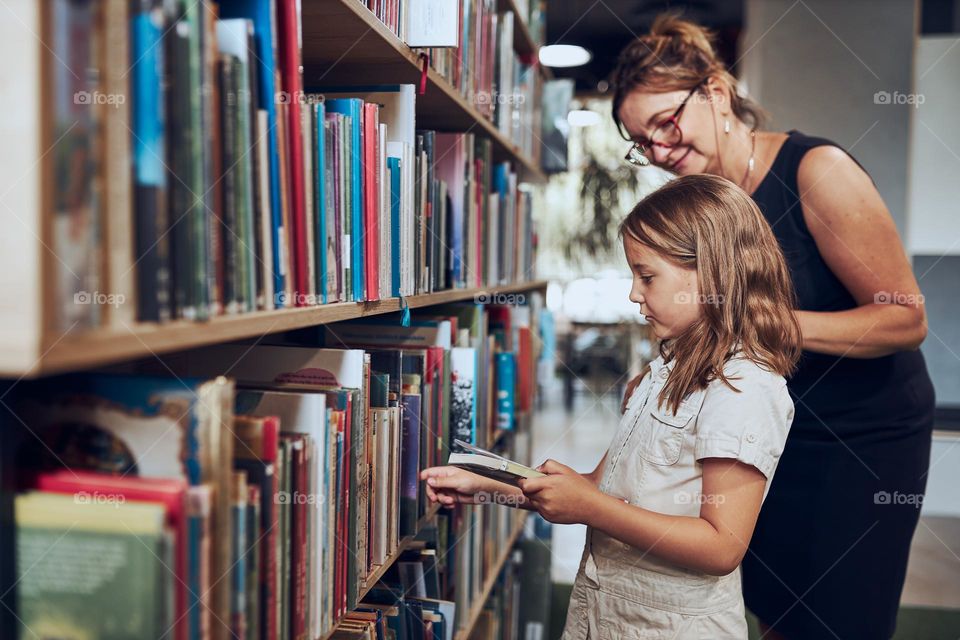 Teacher helping to choose book her schoolgirl in school library. Smart girl selecting literature for reading. Books on shelves in bookstore. Learning from books. School education. Benefits of everyday reading. Child curiosity