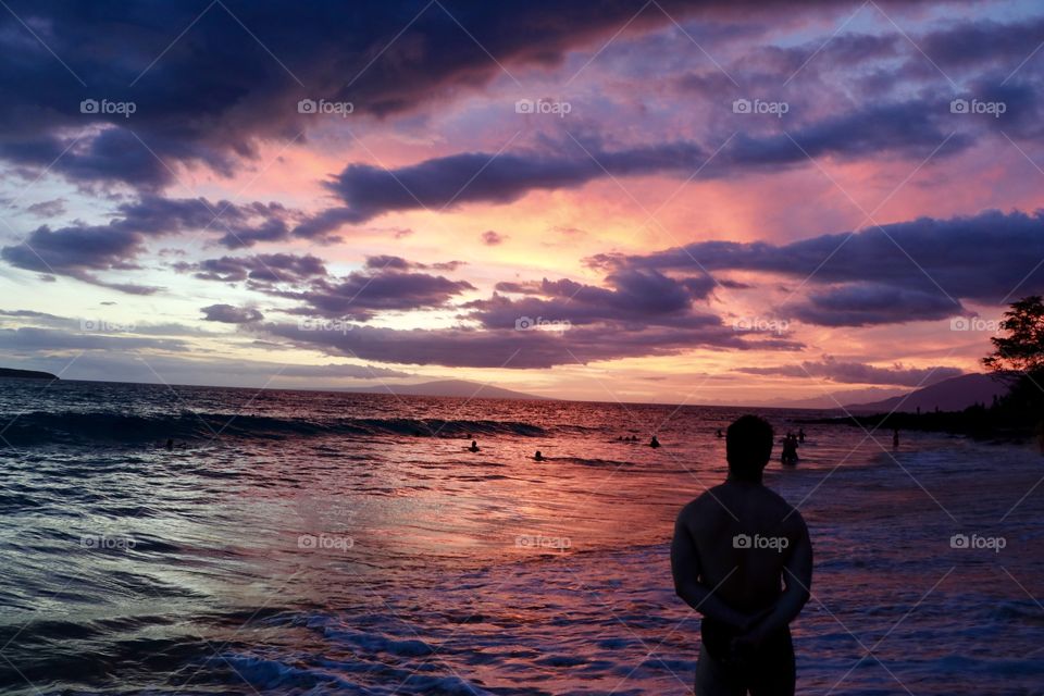 Young man on the beach watching the people  jumping on the waves in the ocean after sun sets in and dusk slowly re-colouring the ocean and the dramatic sky.. 