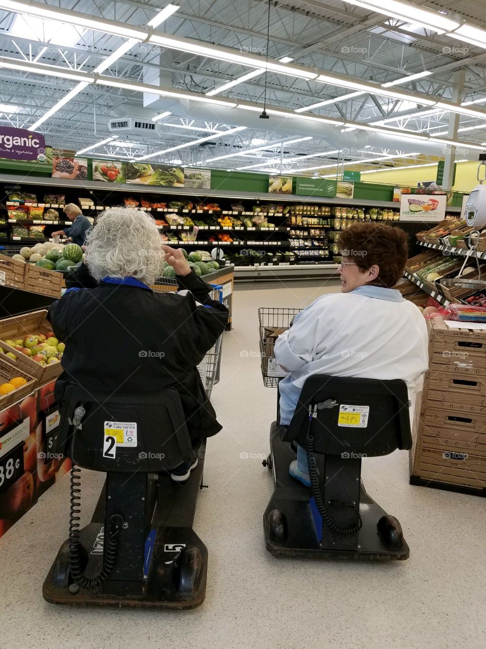 Two older women grocery shopping, both driving motorized handicapped carts as they talked together. They are driving side by side. Ahead is the vegetable department and brightly lit store.