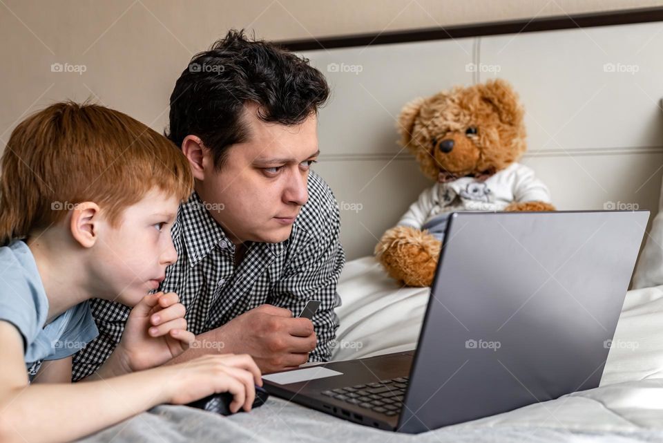 Red-haired boy with his father watching a movie on a laptop lies on the bed