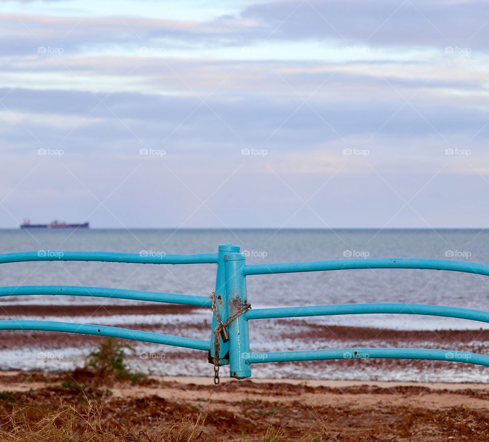 Marine blue metal fence gate with padlock foreground, cargo ship in ocean on the horizon in blurred background 