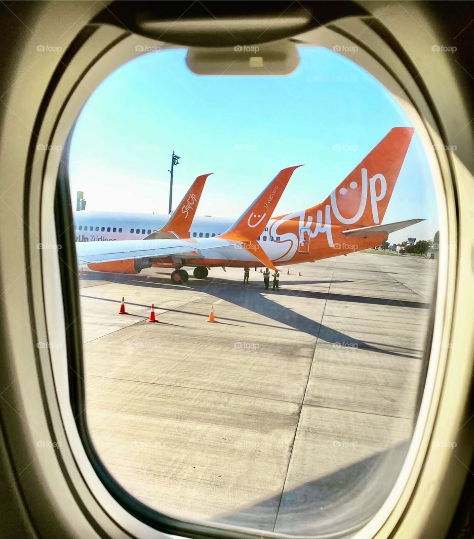 На фото, сделанном из иллюминатора самолета, видно несколько рядом стоящих самолетов. Их корпуса блестят на солнце, а хвосты с логотипами авиакомпаний поднимаются в небо.