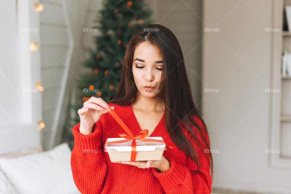 Young beautiful asian woman with dark long hair in cozy red knitted sweater and santa hat with present gift box with red ribbon sitting on bed in the room with Christmas tree