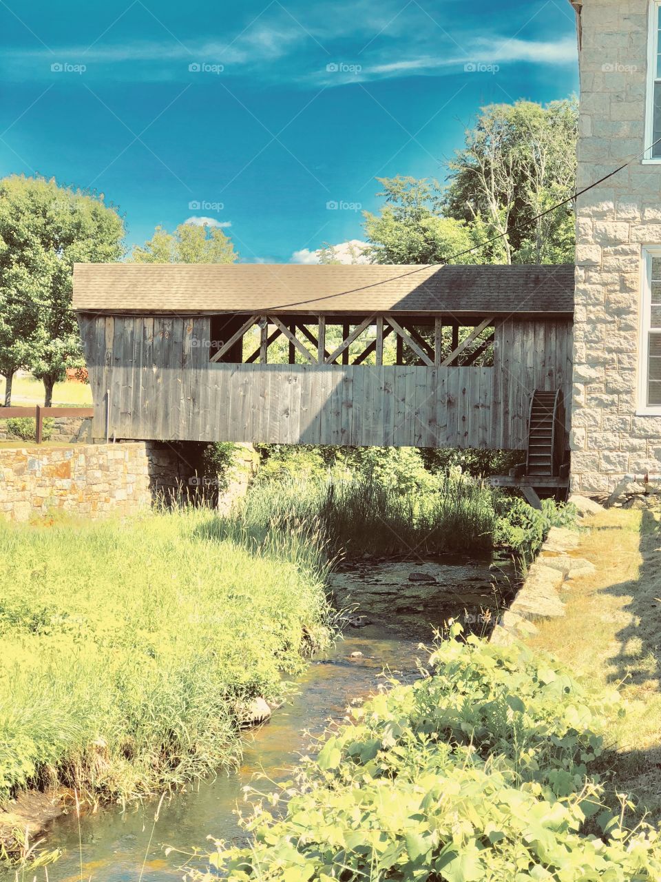 Old mill with water wheel and covered bridge over a stream, grass and trees surrounding it on all sides (Franklin, Massachusetts)