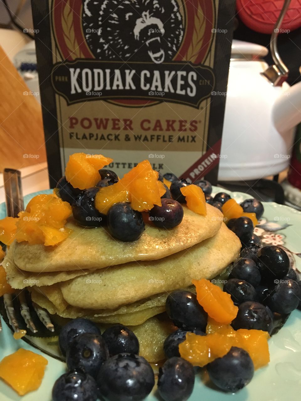 Stack of four Kodiak pancakes with butter, syrup, blueberries and apricots with Kodiak cakes box in background 