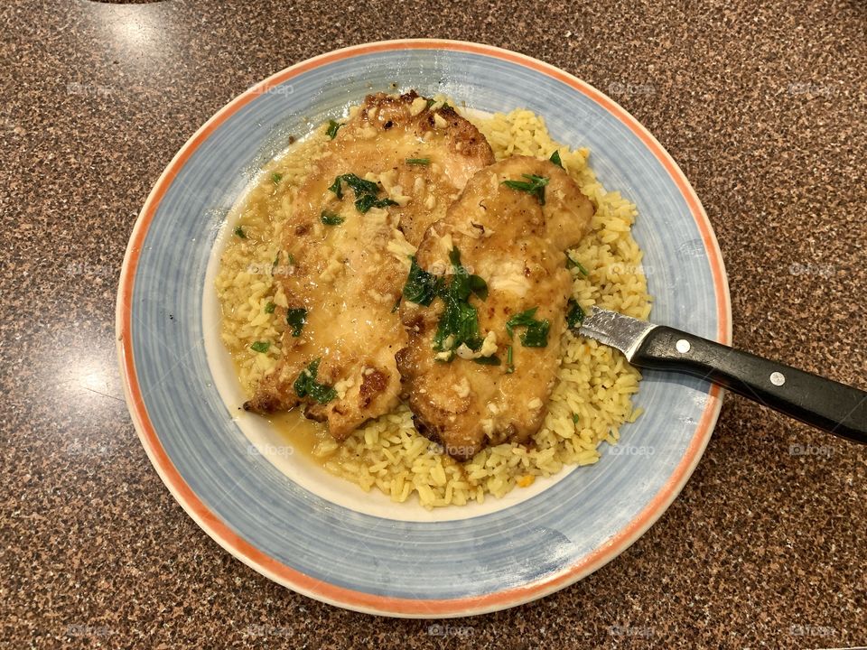 Chicken scampi with garlic sauce, yellow rice for lunch and dinner. Orange and blue round plate. Brown background.