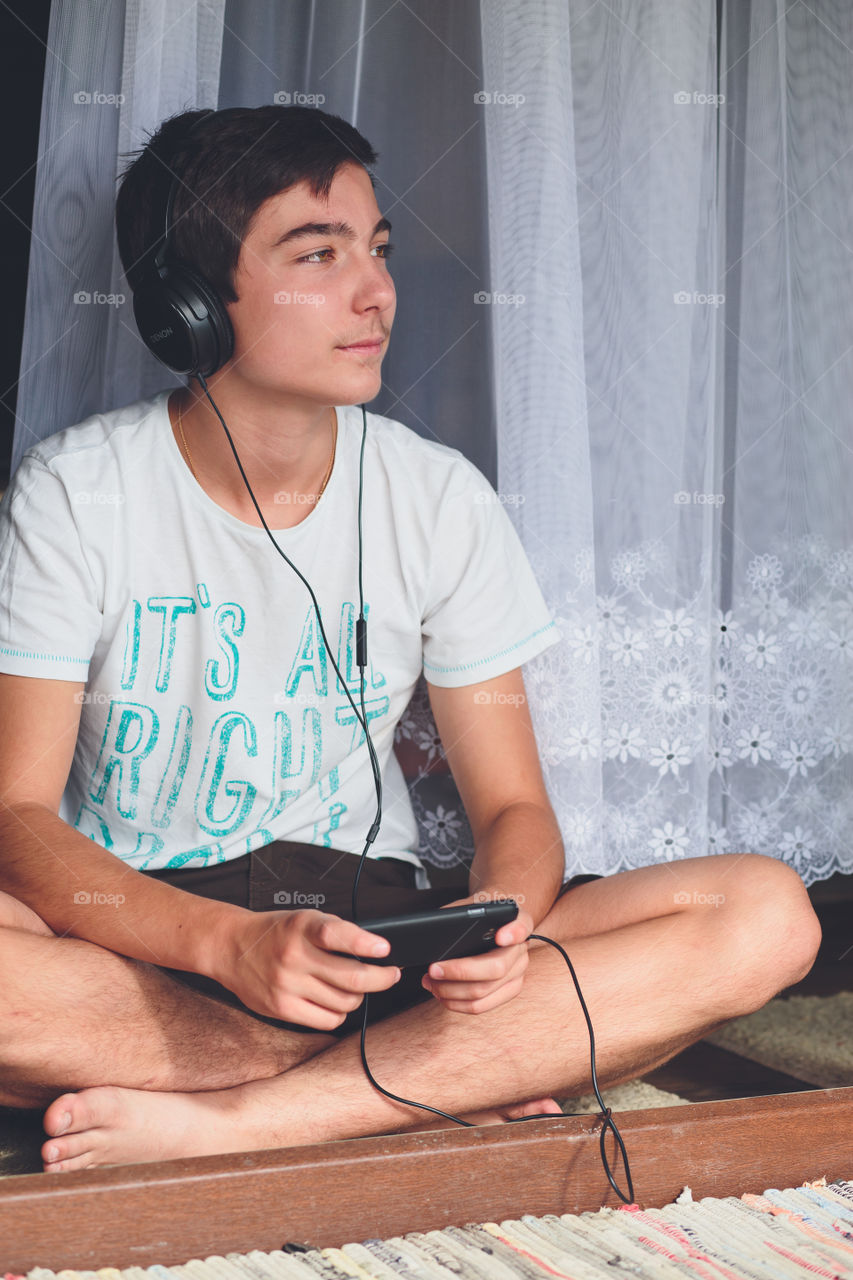 Boy listening to music. Relaxed young boy sitting at the patio door and listening to music from mobile phone