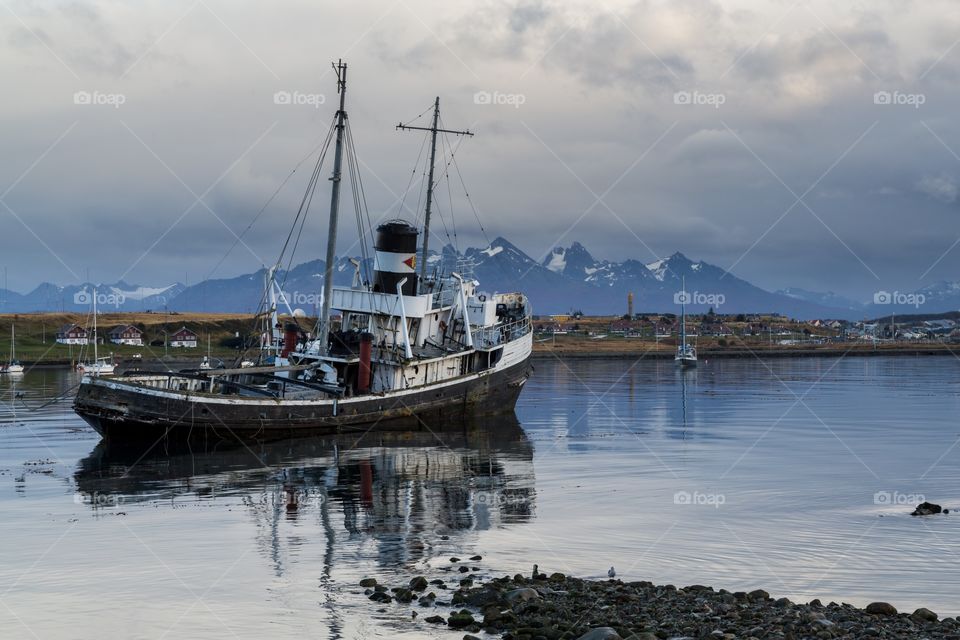 Old boat in mountain scenery. Old boat during sunset facing towards mountains in Ushuaia, Argentina. Cloudy skies. Boat reflection in the water.