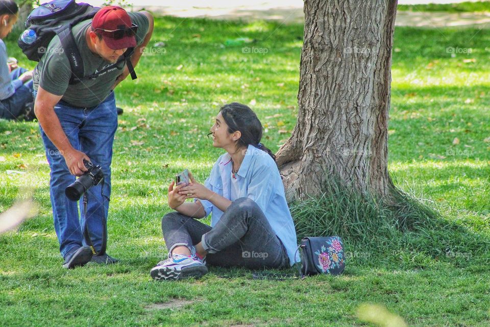 a girl in jeans and a shirt sits on a lawn in a park with a phone in her hands