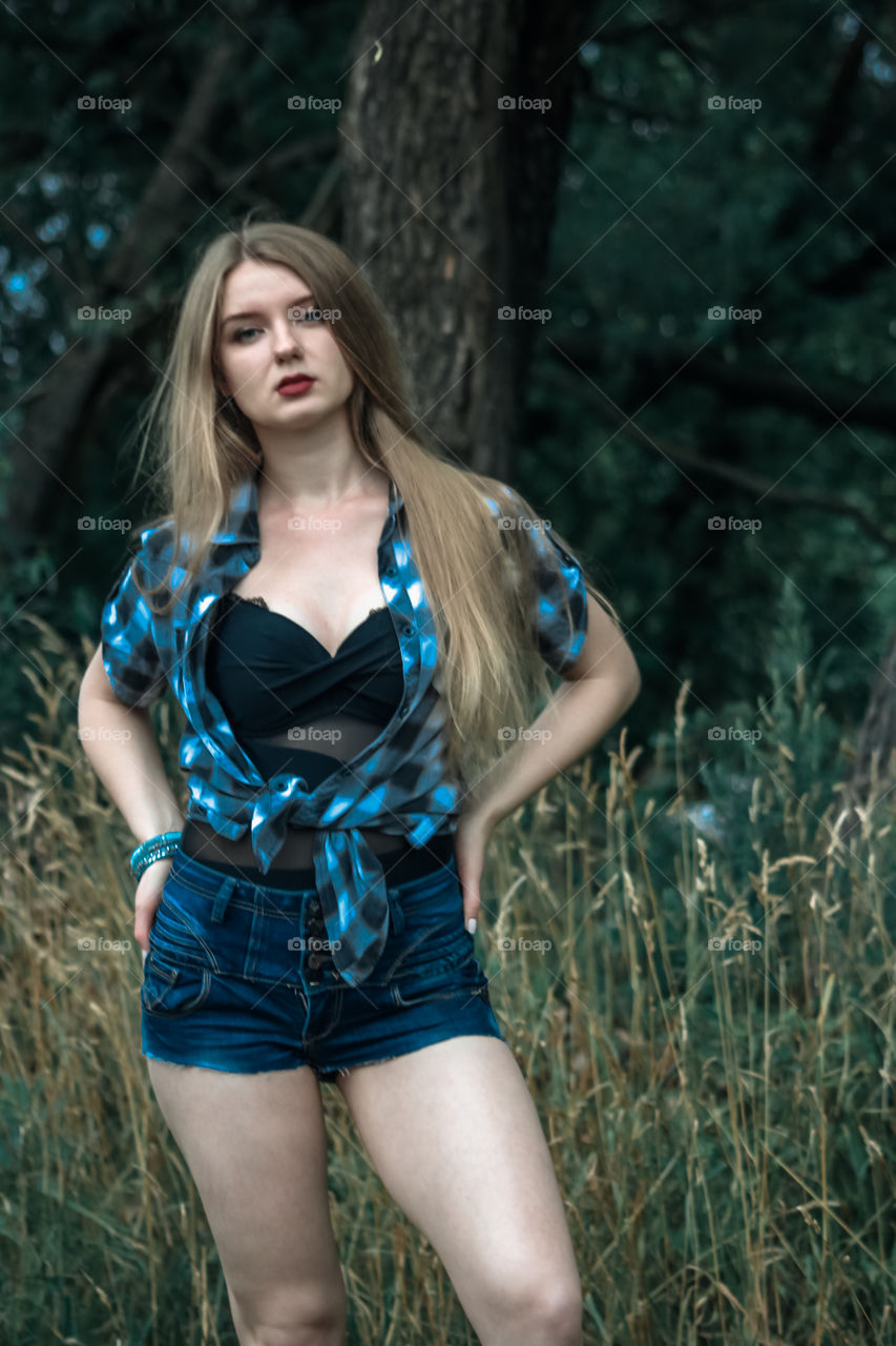 A girl with blond hair in a plaid shirt and short denim shorts on a background of trees and nature Girl, woman, man, people, blonde, blonde hair, checkered shirt, shorts shorts, denim shorts, forest nature, trees, grass, feelings, emotions tenderness, love, lifestyle, lifestyle, recreation