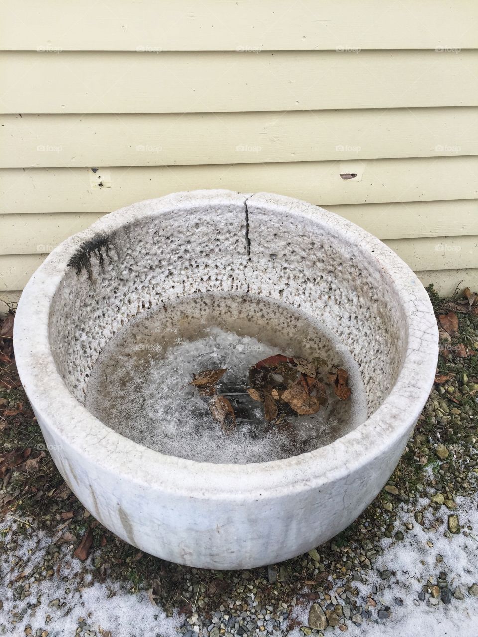 A large, decorative cement bowl partially filled with ice and leaves. It sits by an exterior wall on gravel.