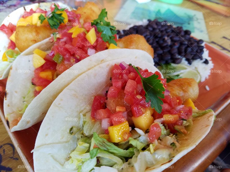 Fish tacos on flour tortilla topped with mango salsa with a side of white rice and black beans.