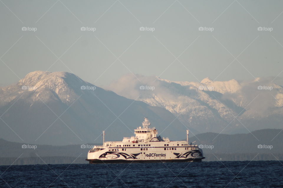 Shot of a BC ferry crossing the strait lit with the rare winter sunlight. The sea is indigo blue, the hills behind dark green,  the mountains capped with fresh snow and small wisps of powdery clouds kiss the mountain peaks. 