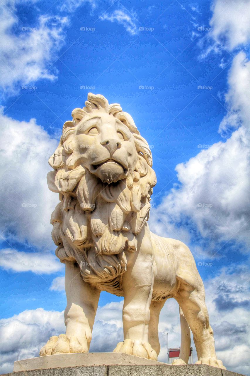 Proud British Lion. A stone lion statue proudly standing on a plinth with a blue, cloudy sky behind.
