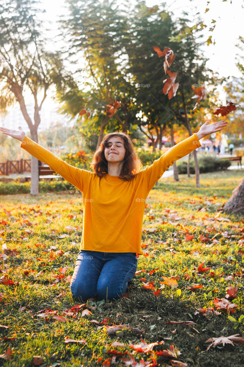 Pretty teenager girl with yellow sweater throwing up dry leaves sitting at autumn grass 