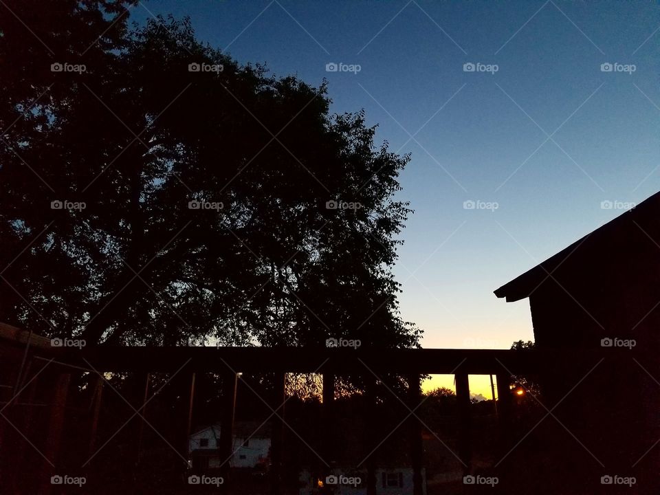 Sunset fades from yellow to dark blue with a tree, house and fence in silhouette