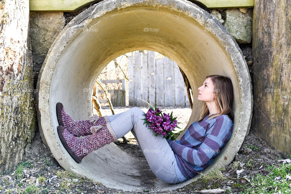 Millennial woman sitting in the opening of a cement tunnel holding a bouquet of flowers 