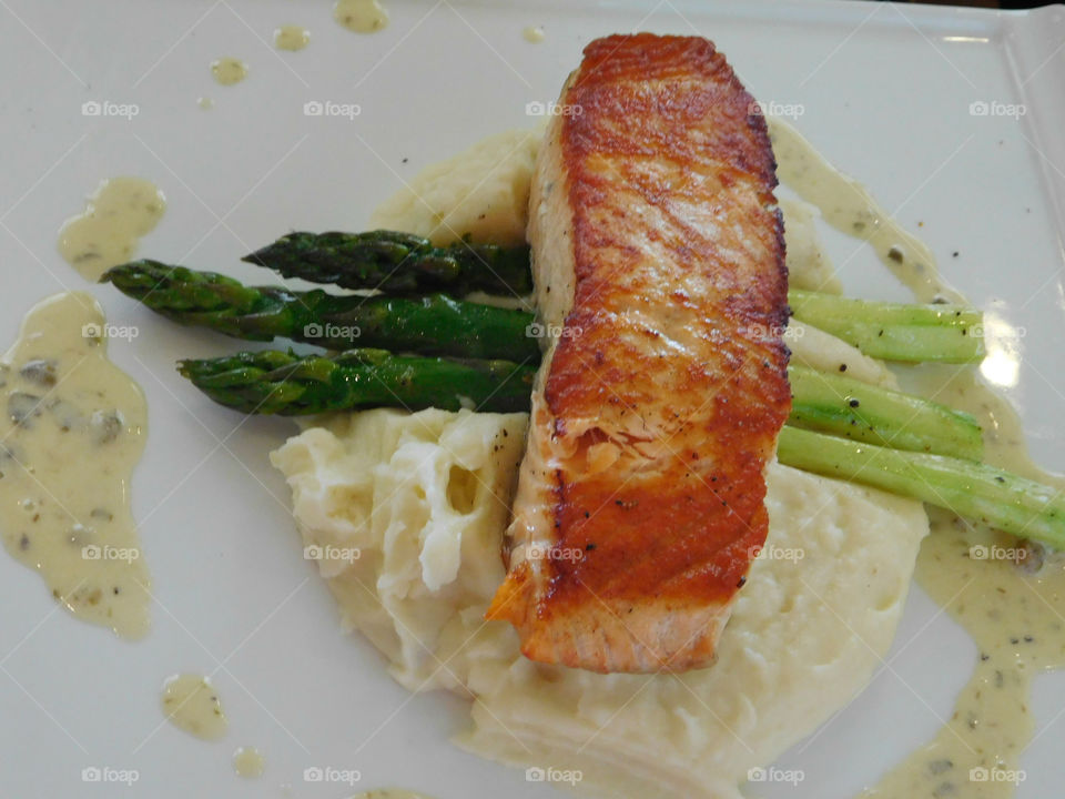 A fresh salmon steak on a bed of seasoned mash potatoes and crispy asparagus lined with tarter sauce sprinkled on the plate! So flaky it will melt in your mouth!