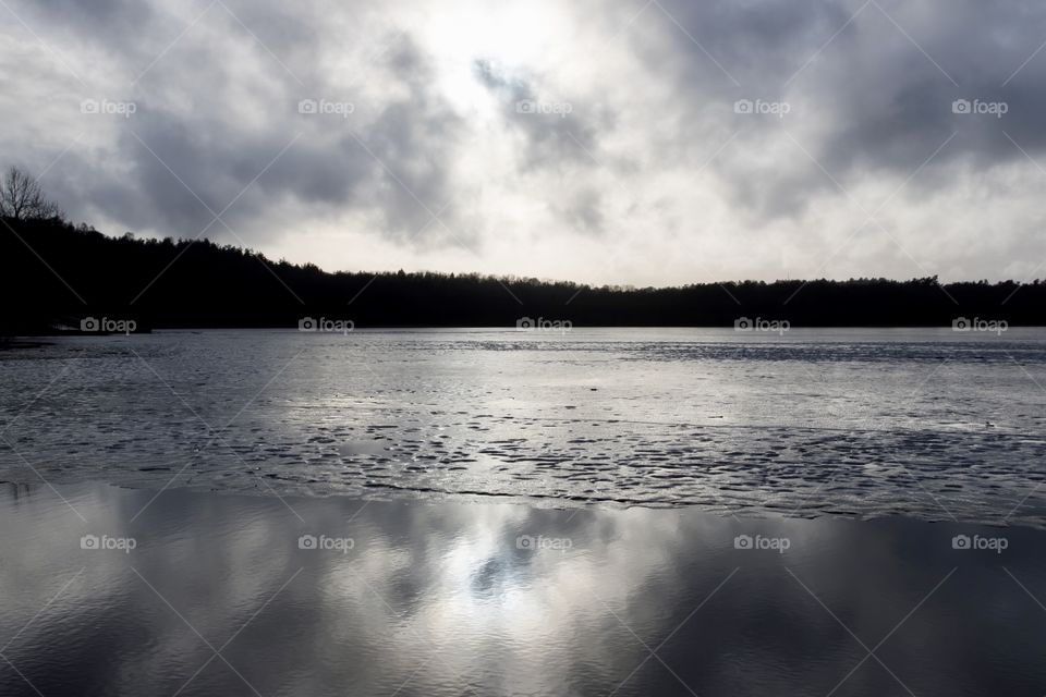 A dark grey day with dramatic clouds and reflection on the water. The ice on the lake is melting. Dramatiska moln och reflektion på sjön. Isen börjar smälta. Härlanda tjärn Sverige