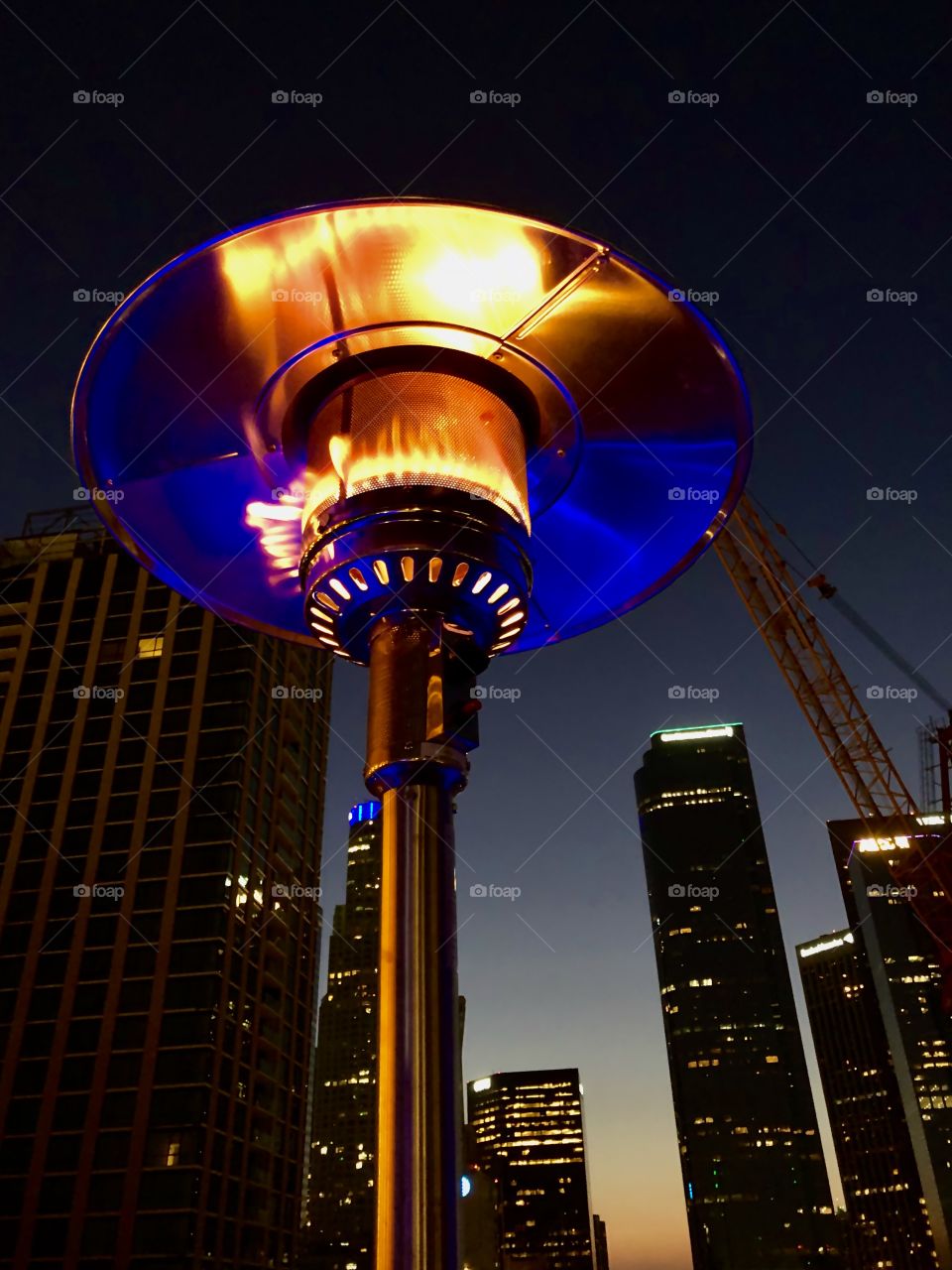 Blue lights from a railing below reflect up into the hood of a heatlamp with flame; a downtown Los Angeles rooftop on a chilly summer evening. 