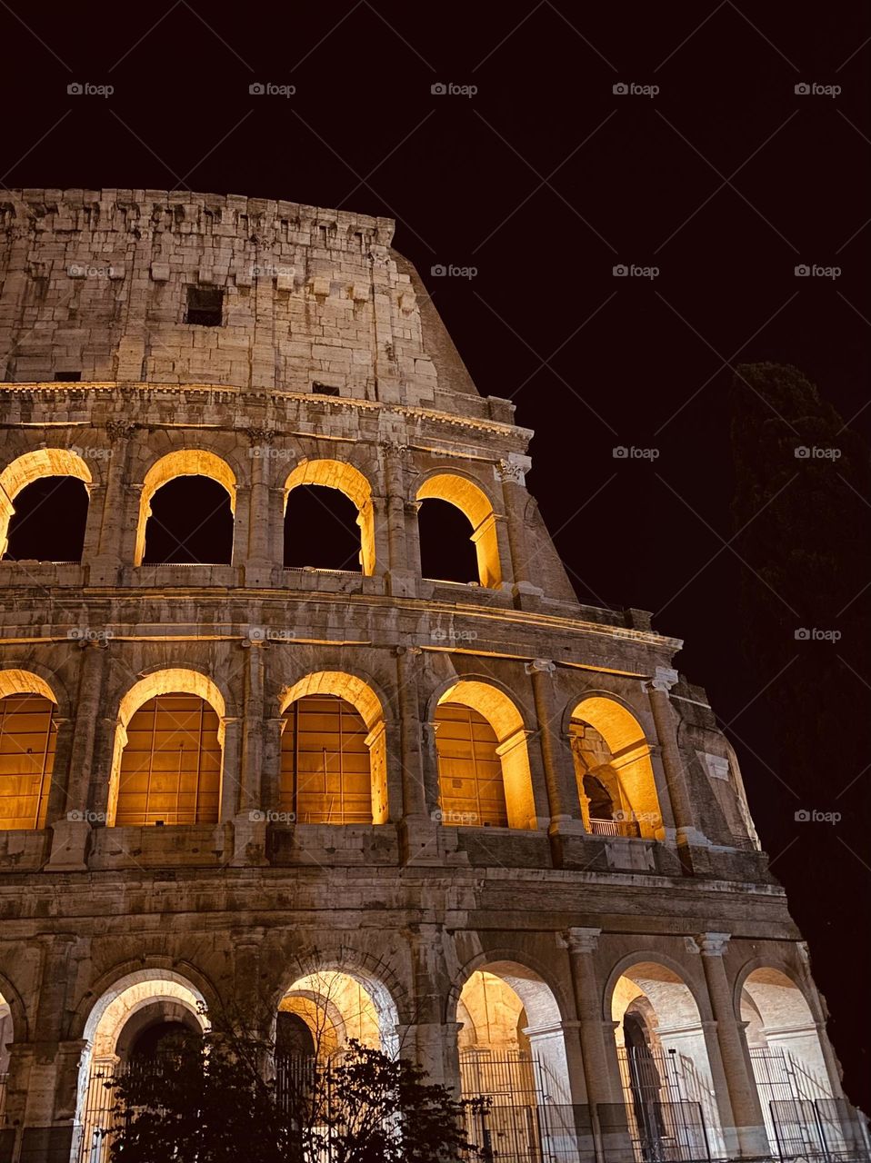 The Colosseum rises majestically under the night sky, its ancient arches glowing with warm golden light. Shadows accentuate the weathered stone, telling timeless stories of Rome's grandeur.