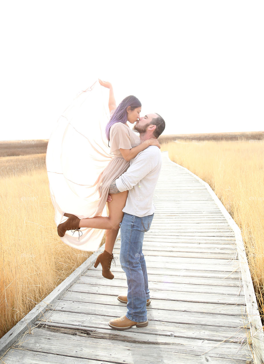 Young adult couple in love posed outdoors wooden plank walkway with golden wheat straw grass. Man casually in denim blue jeans, brown cowboy boots, off-white long sleeved sweater. Woman long hair purple brown boots, mauve dress, outstretched arm