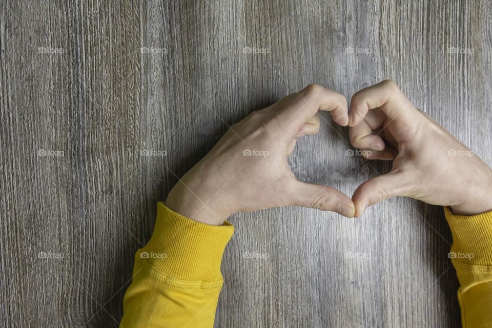 Men's hands folded in the shape of a heart in a yellow jacket lie on a gray wooden surface, which is used as a background or a surface with incident light.