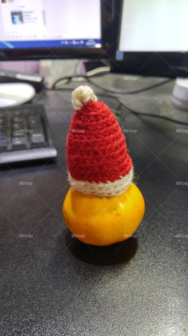 A funny looking tangerine with a santa hat on an office desk.