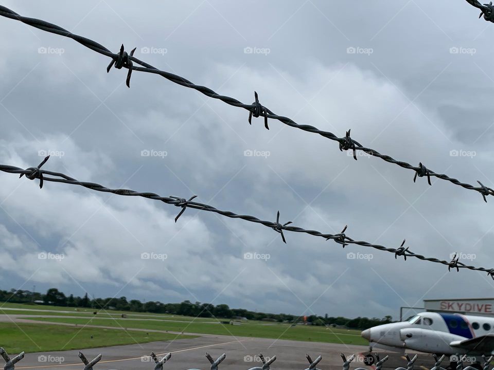 Small Airport for leisure and activities and other uses and purposes for outdoor fun and skydiving. Seeing a plane by the plane track behind a link chain fence with anti climb barrier on top with sharp barbed wire during a cloudy day.