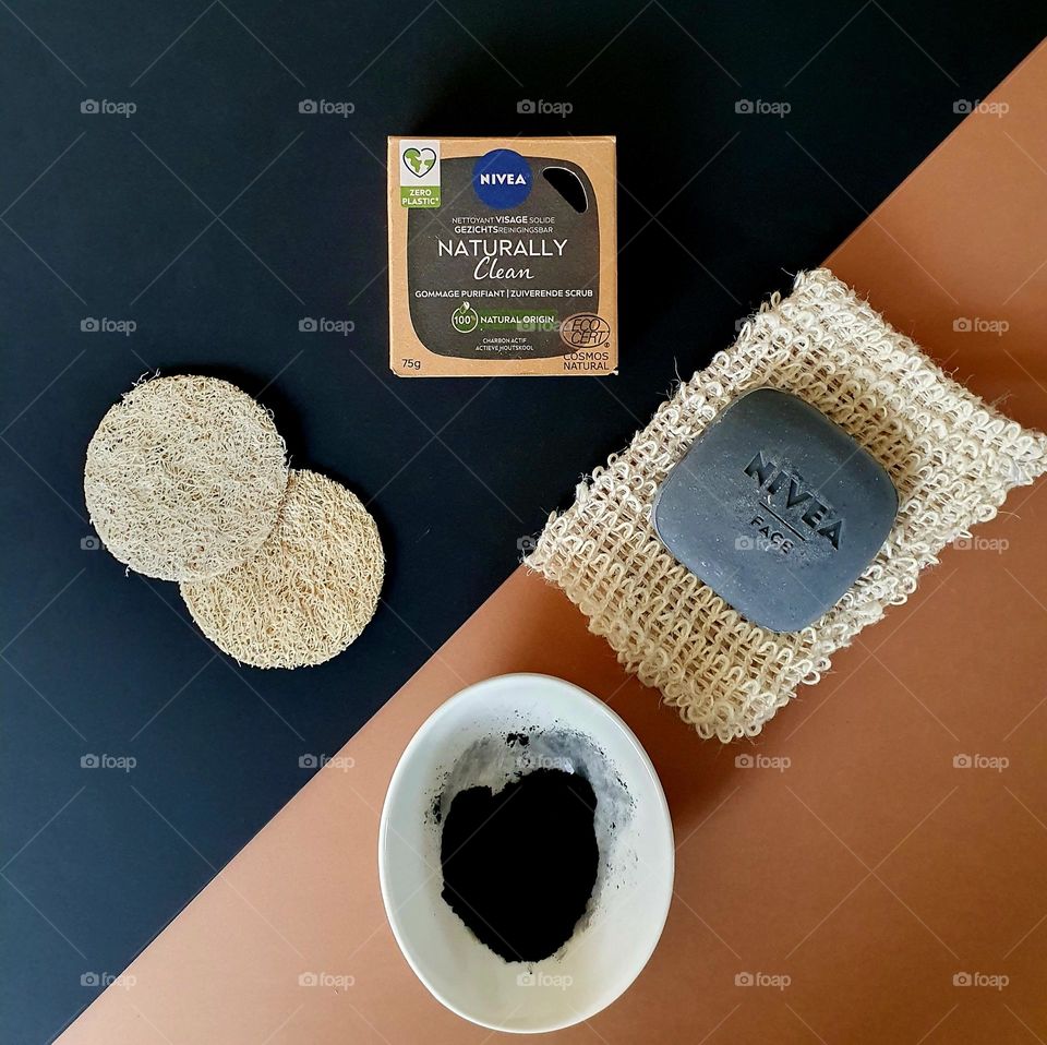 Flatlay Nivea Naturally Clean purifying scrub soap placed on black and brown paper, and composed of a china bone little bowl filled with charcoal power, the Nivea soap and its box, two scrubbing pads and a natural fibre soap pouch
