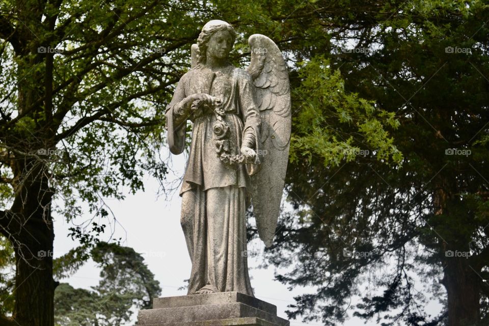 A grey, weathered, concrete sculpture of an Angel wearing multi layered clothing holding a string of flowers and having bare feet