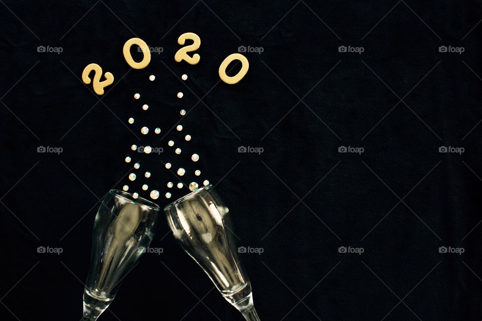 Flat lay of the numbers ‘2020’ in gold above gold-tinted champagne glasses with rhinestone bubbles on black background - copy space on right side of landscape format