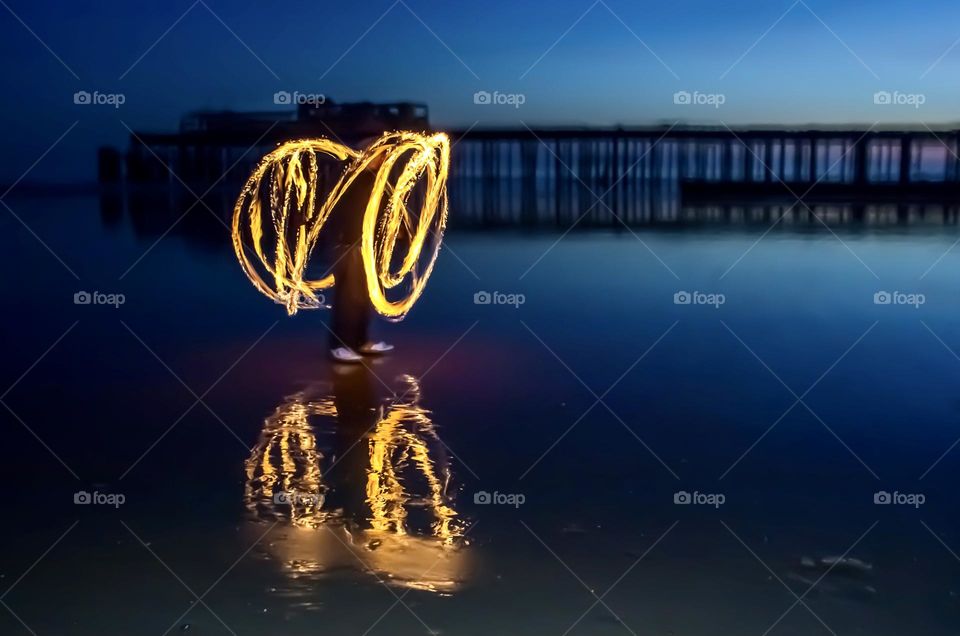 A person spinning fire poi on a wet beach, in front of a pier