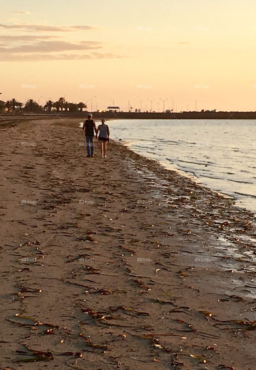 Senior couple holding hands walking along beach toward sunrise 