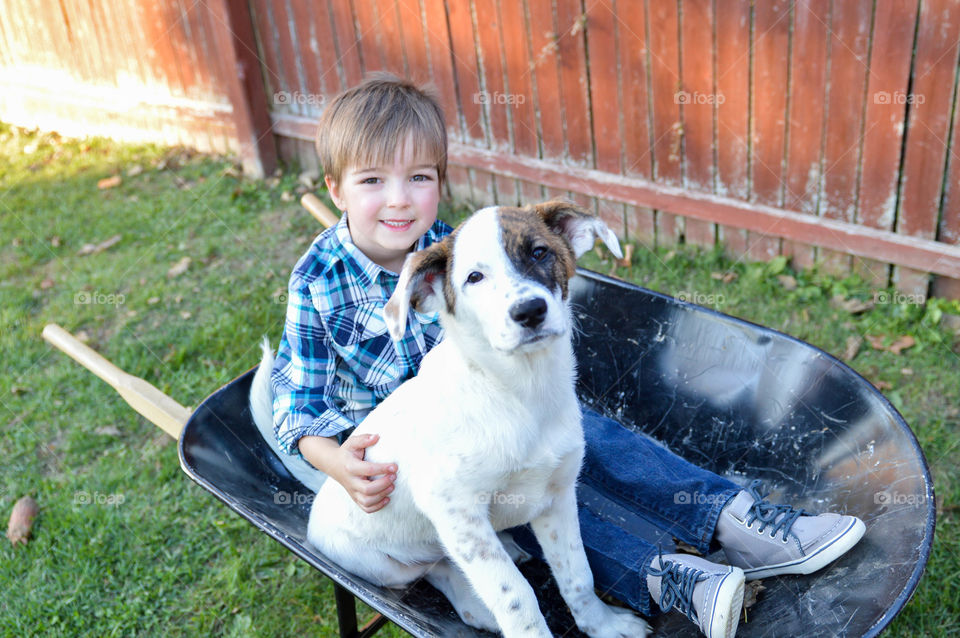 Young boy and mixed breed puppy sitting in a wheelbarrow outdoors