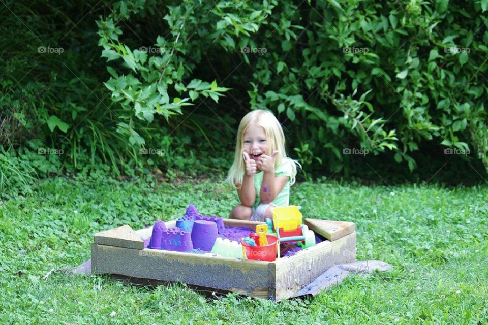 Little girl giving two thumbs up to her new purple sand for her sandbox 