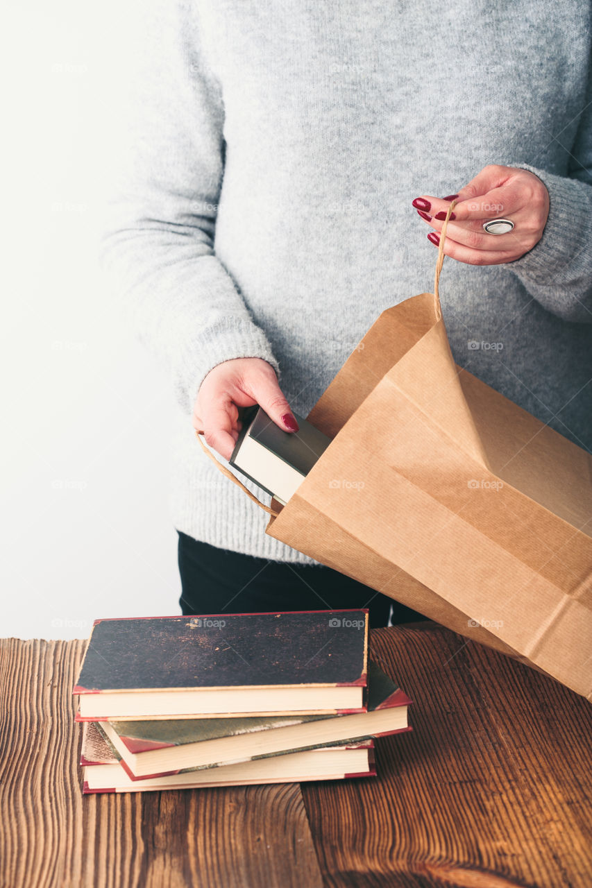 Young woman putting old books to paper bag in antique bookstore. Woman wearing grey sweater and jeans. Vertical photo