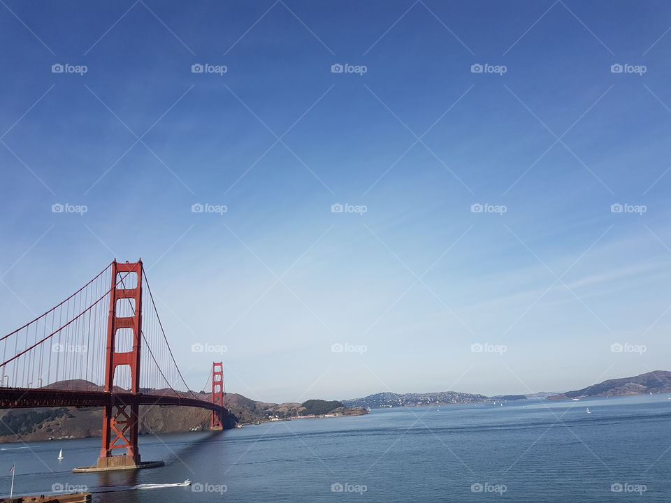 side view from land of red colored Golden Gate Bridge and blue ocean bay in San Francisco, America, on cloudy blue sky day