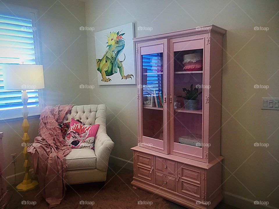 A little girls bedroom with a rectangular pink painted dresser with squares shaped glass windows a sitting chair, lamp and natural sunlight. USA, America