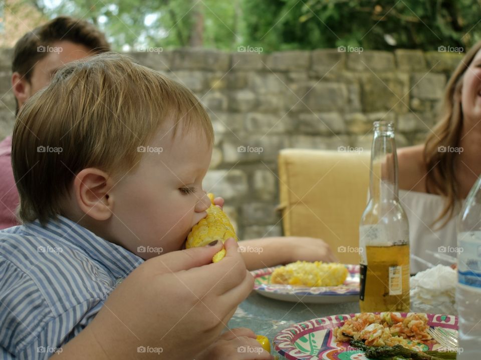 Cute baby boy eating corn from the cob at backyard BBQ party