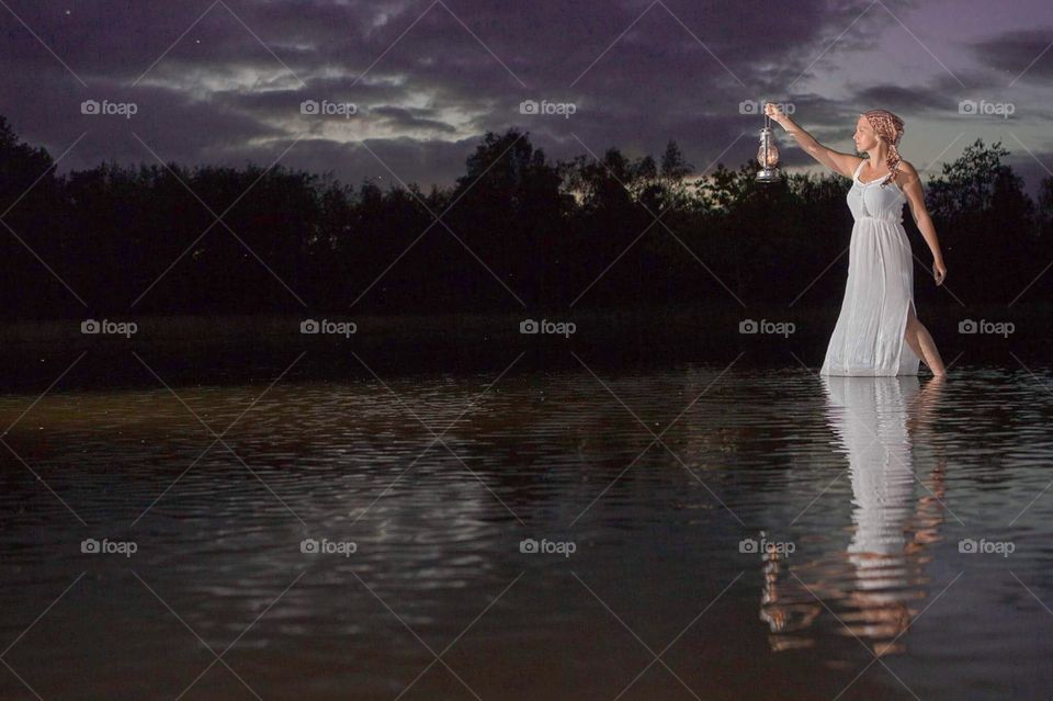 Beautiful young blonde woman standing in the water of a lake around Dusk, sunset, wearing a long white dress and holding a vintage lamp