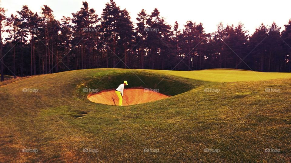 Beautiful view on golf player playing from the bunker (sandy area) with trees and green behind. With daylight.