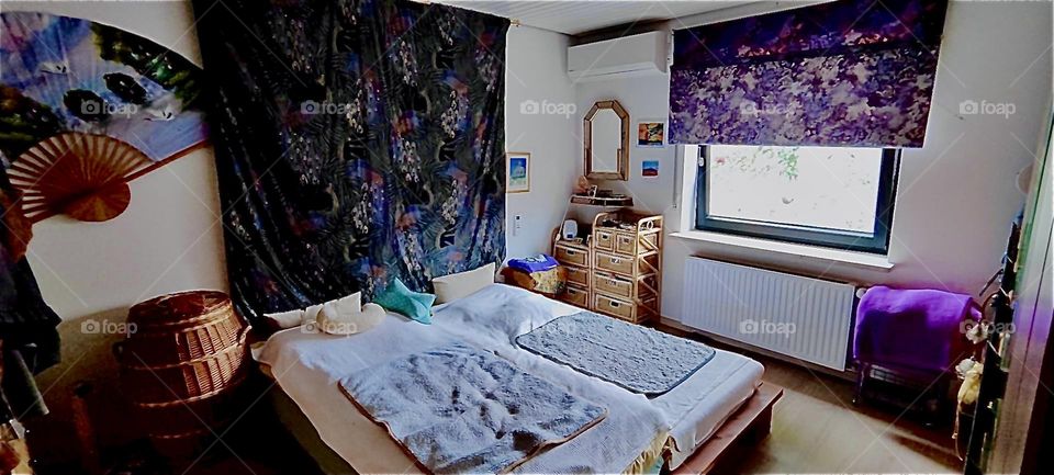 This is the bedroom in the lovely bungalow in “Bavaria”, Germany. A big ornamented tapestry hangs from the wall behind the bed. Matching curtains, a mirror, a wicker rattan chest with drawers  complete the interior design. 2023. Hypnotic Productions